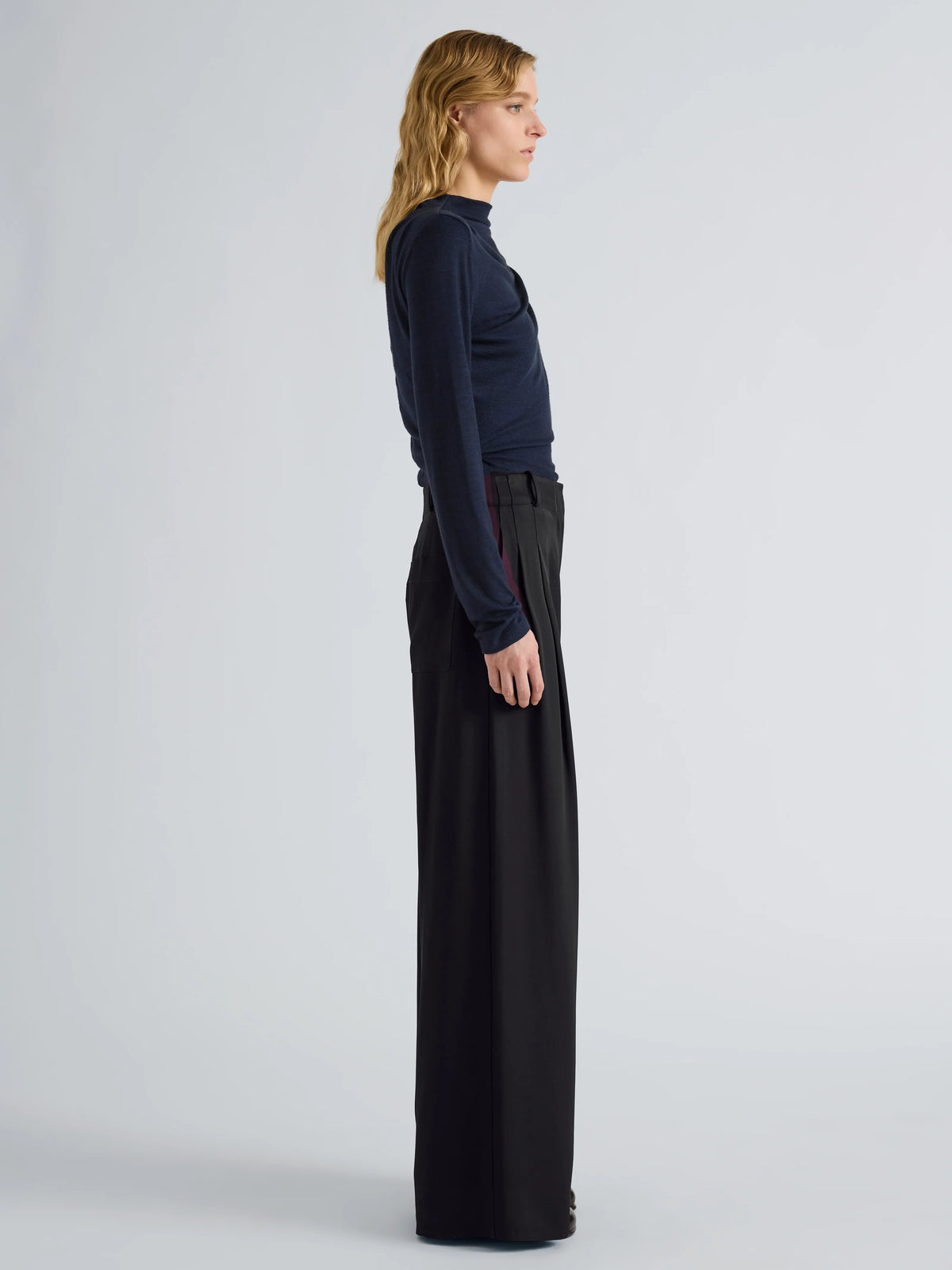 A woman stands in profile wearing a dark navy long-sleeve mock-neck top and black wide-leg trousers against a light gray studio background.