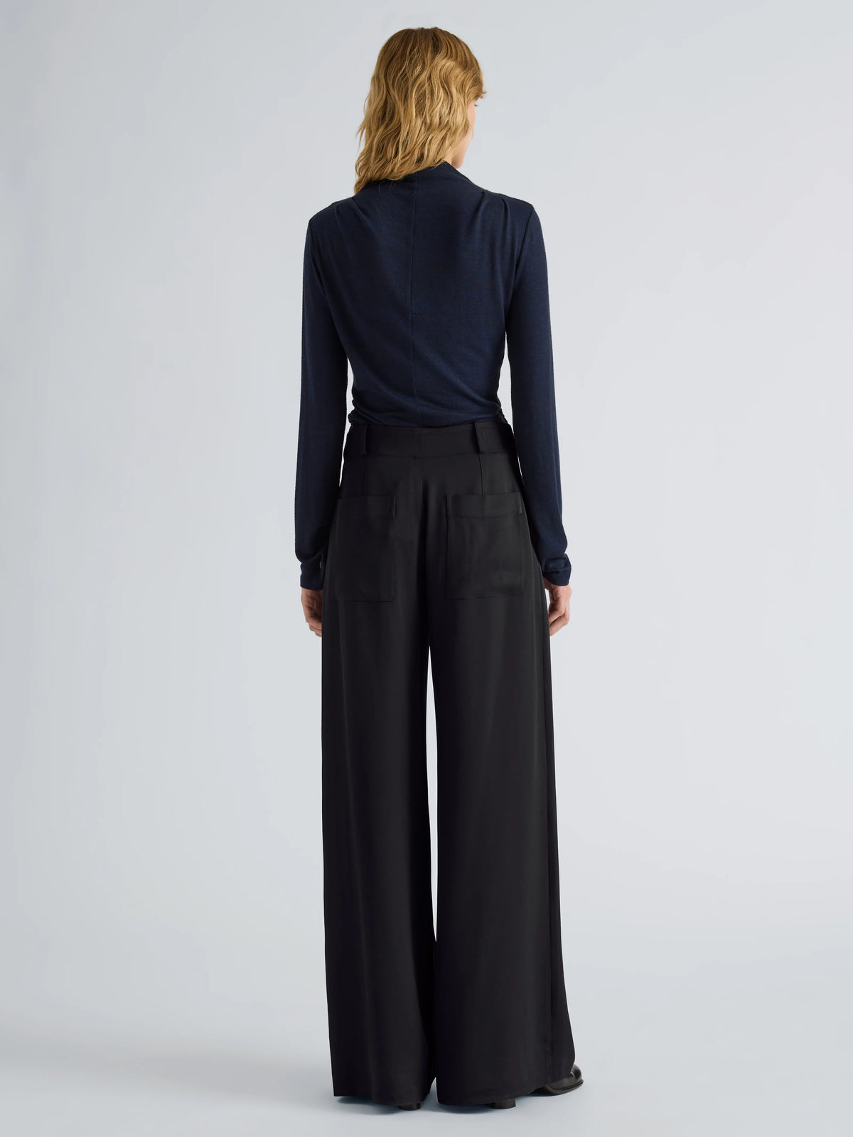 A woman viewed from the back wearing a dark navy long-sleeve mock-neck top and black wide-leg trousers, standing against a light gray studio background.