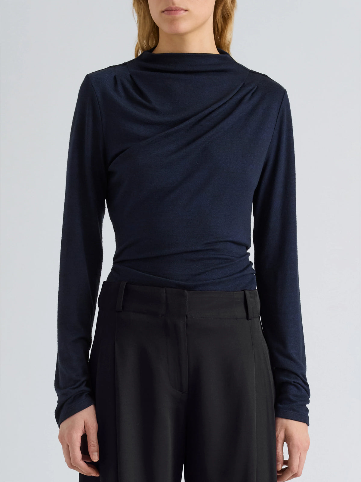 A model wearing a dark navy blue long-sleeve top with a draped, asymmetrical cowl neckline, paired with black trousers, against a light gray studio background.