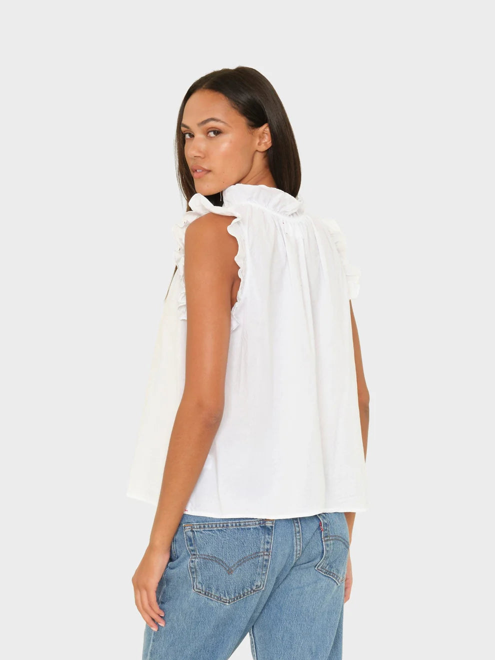 Woman wearing a white sleeveless top with ruffle details and blue jeans on a light gray background