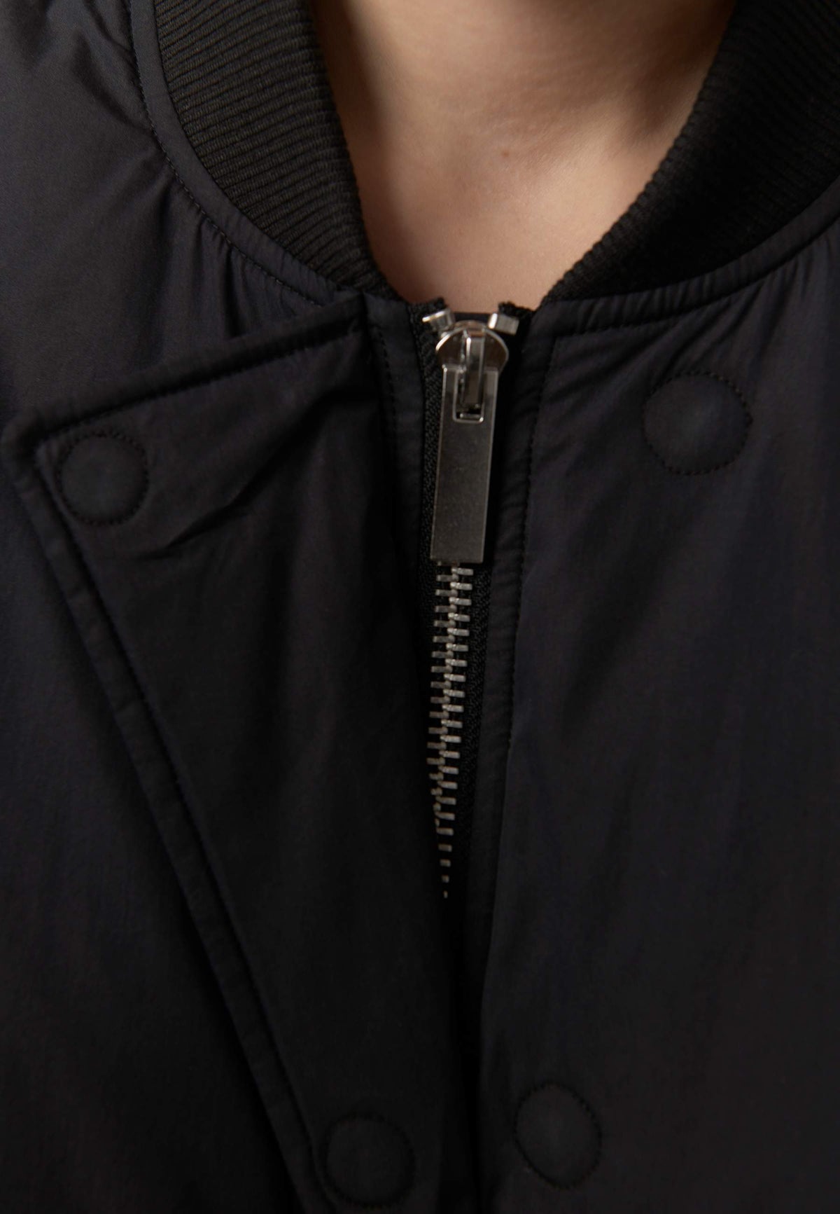 Close-up of a black bomber jacket featuring a central silver zipper, snap-button closure, ribbed collar, and a model's neck visible at the top.