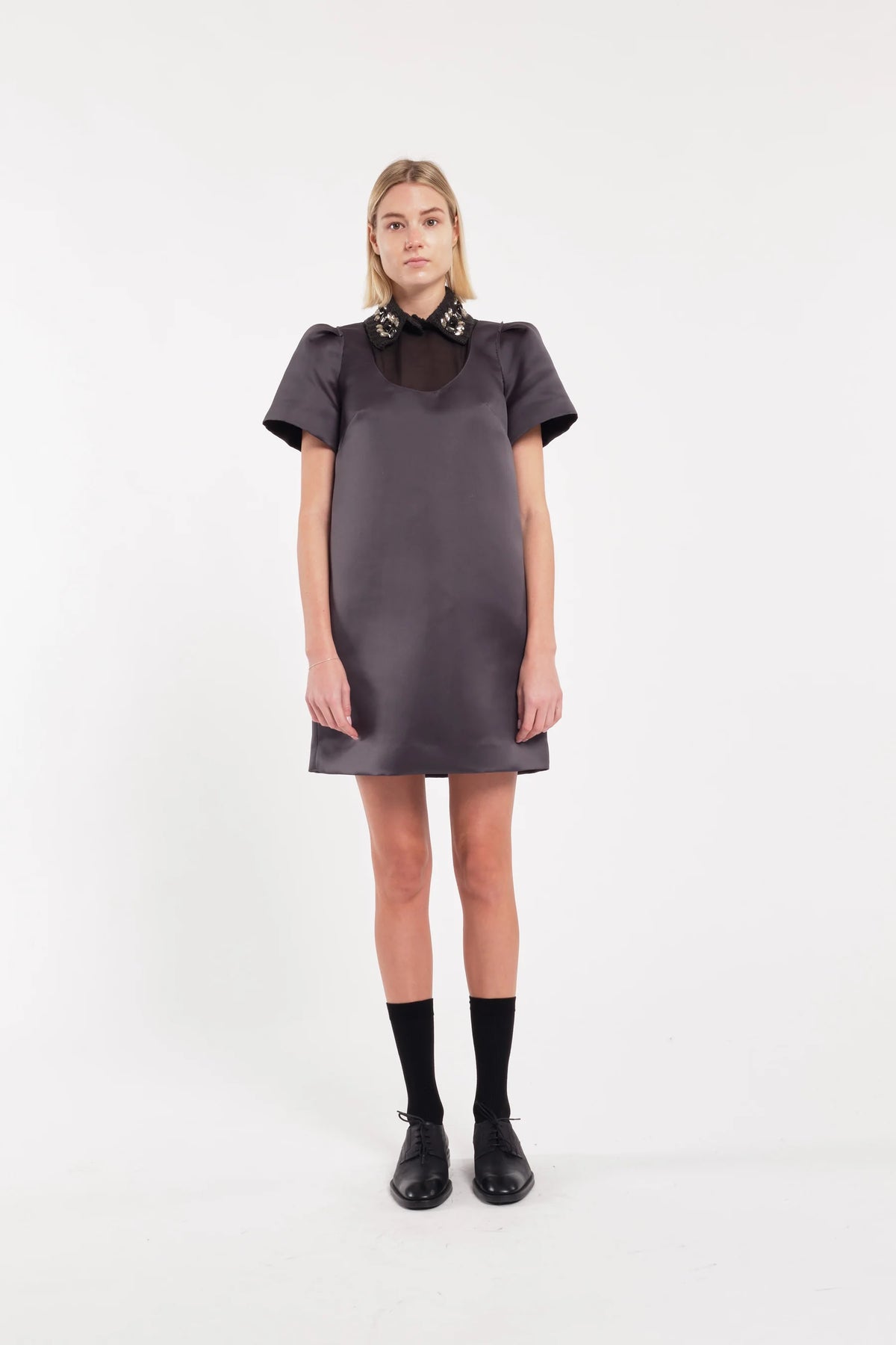 A model stands against a plain white studio background wearing a dark gray short-sleeve satin dress with a contrasting dark brown collar, black socks, and black leather shoes.