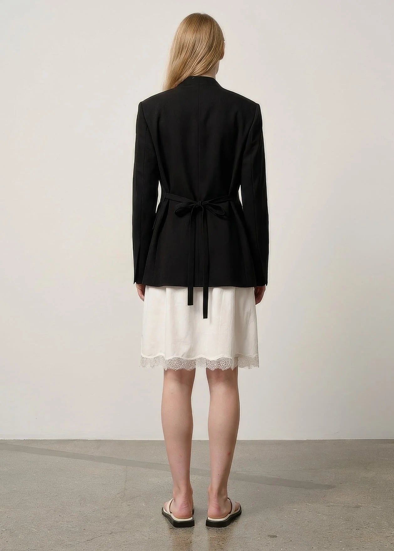 A woman viewed from the back wearing a black long-sleeve wrap-style blazer with a tie waist over an off-white skirt, standing against a plain light wall.