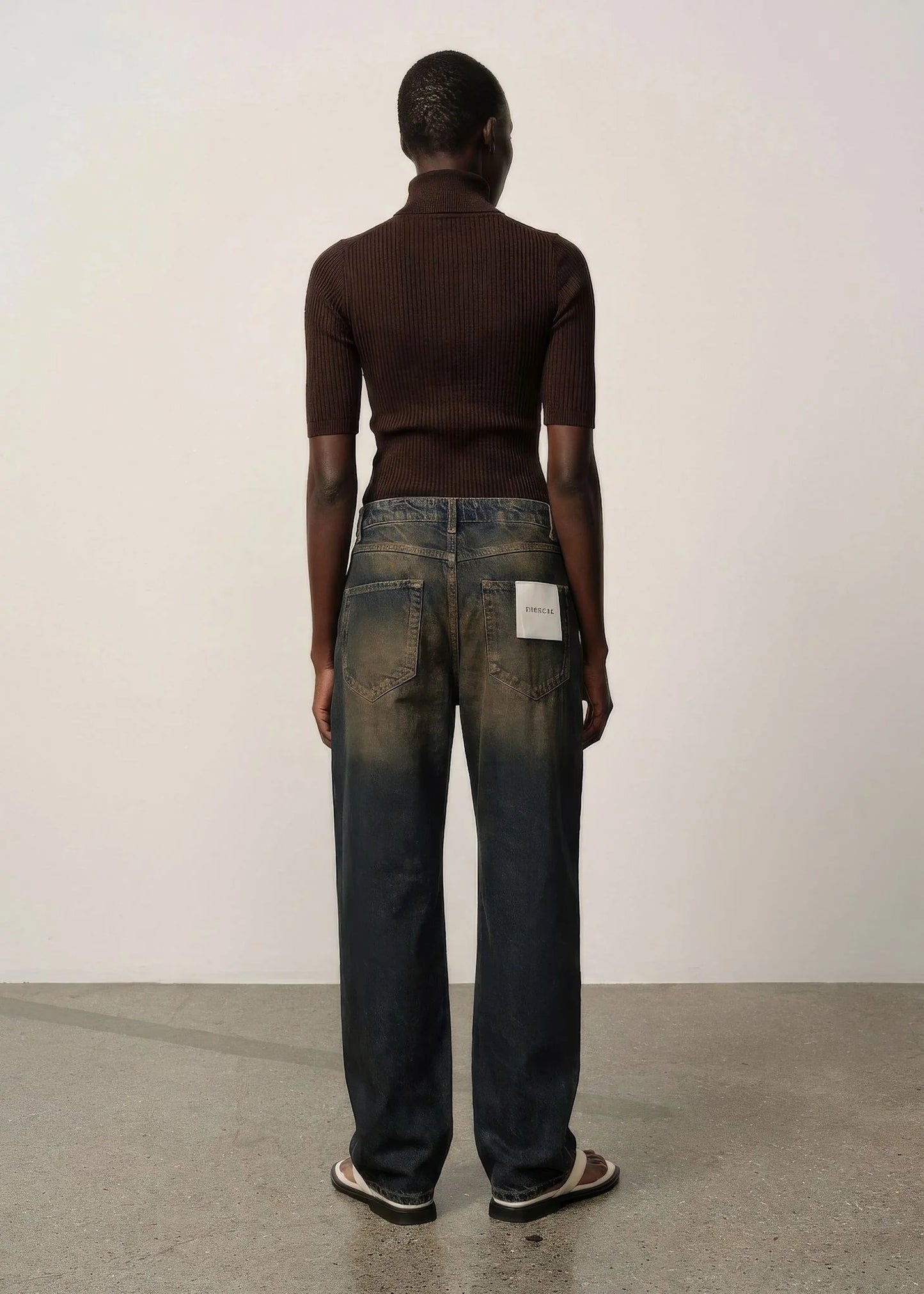 Person wearing a brown turtleneck and blue jeans standing against a white wall.