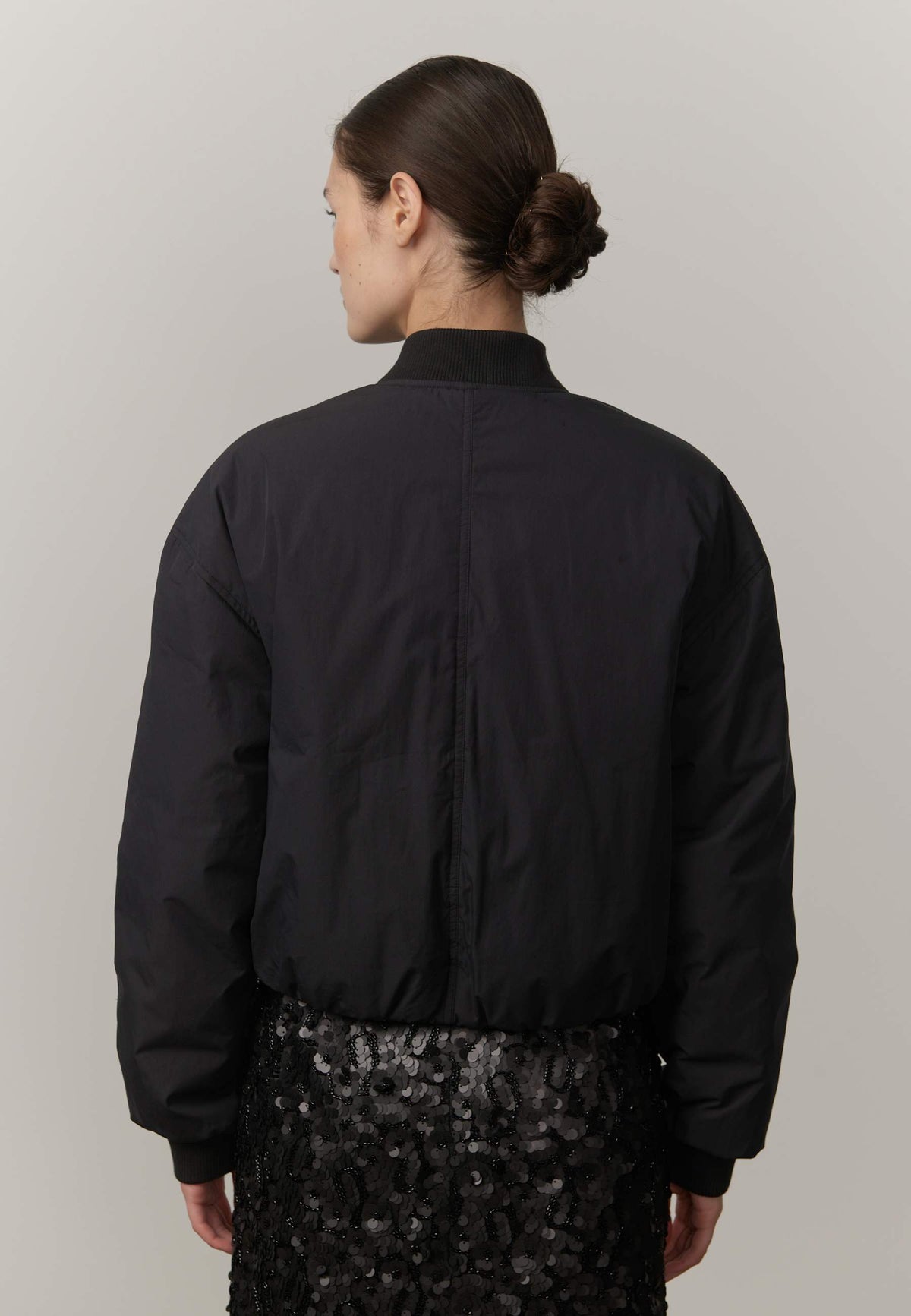 A woman shown from the back wearing a black bomber jacket and a black sequined skirt against a plain light gray studio background.